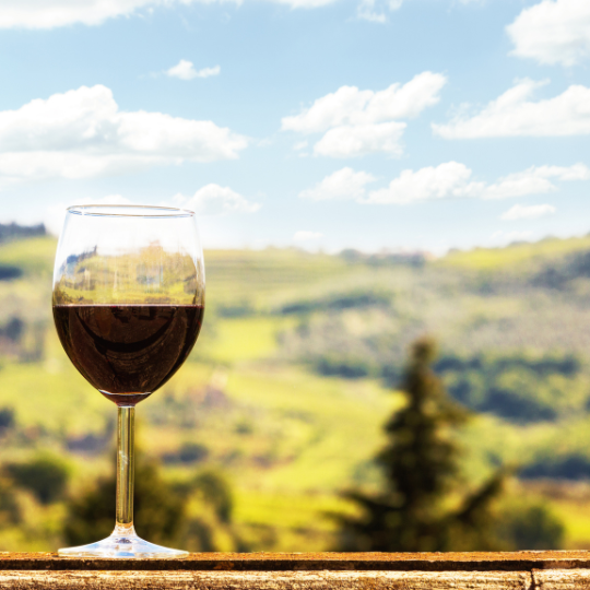 Toda comida italiana debe ir acompañada de un delicioso vino. En primer plano, una copa llena de delicioso vino tinto italiano. Al fondo, un paisaje típico italiano.