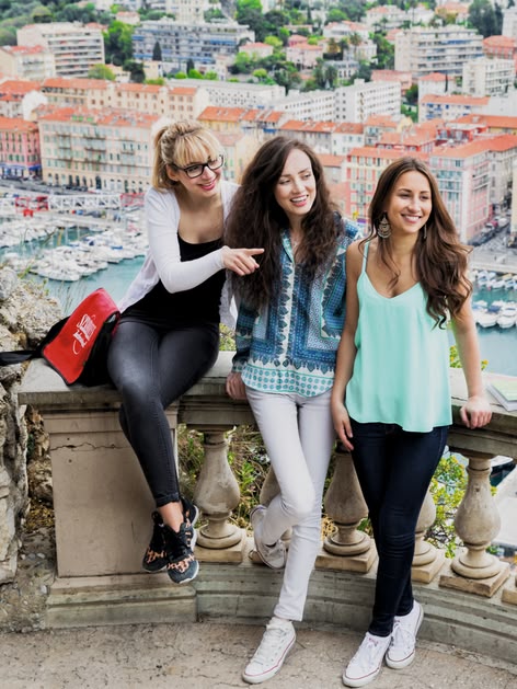 Drie Sprachcaffe-studenten glimlachen op een balkon met uitzicht op Nice, Frankrijk, met de haven en kleurrijke gebouwen rondom