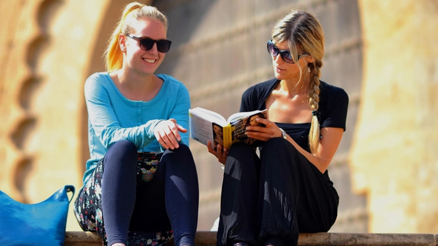 Twee vrouwen zitten op een stenen trap een boek te lezen in Rabat, Marokko, met historische architectuur achter hen.