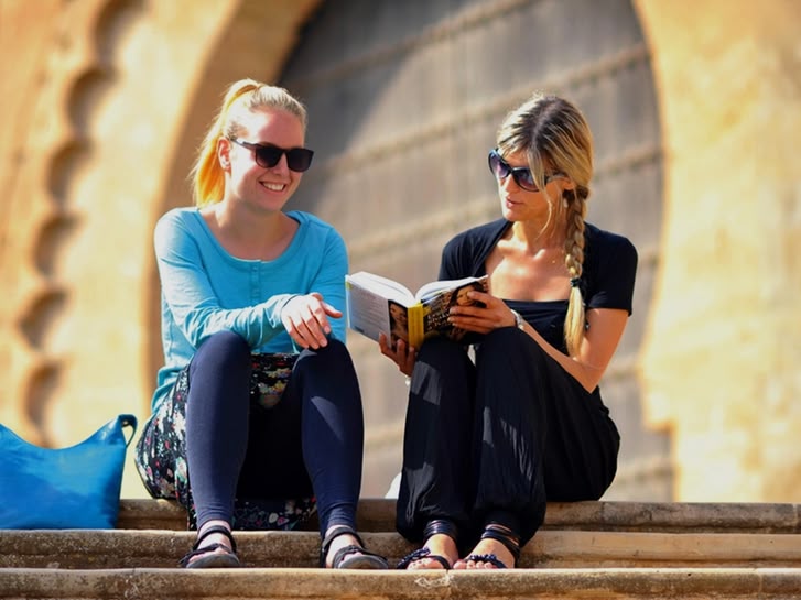 Twee vrouwen zitten op een stenen trap een boek te lezen in Rabat, Marokko, met historische architectuur achter hen.