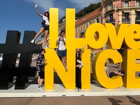 Groep jongeren poseert op en rond het grote gele "#ILoveNICE" bord in Nice, Frankrijk, op een zonnige dag.