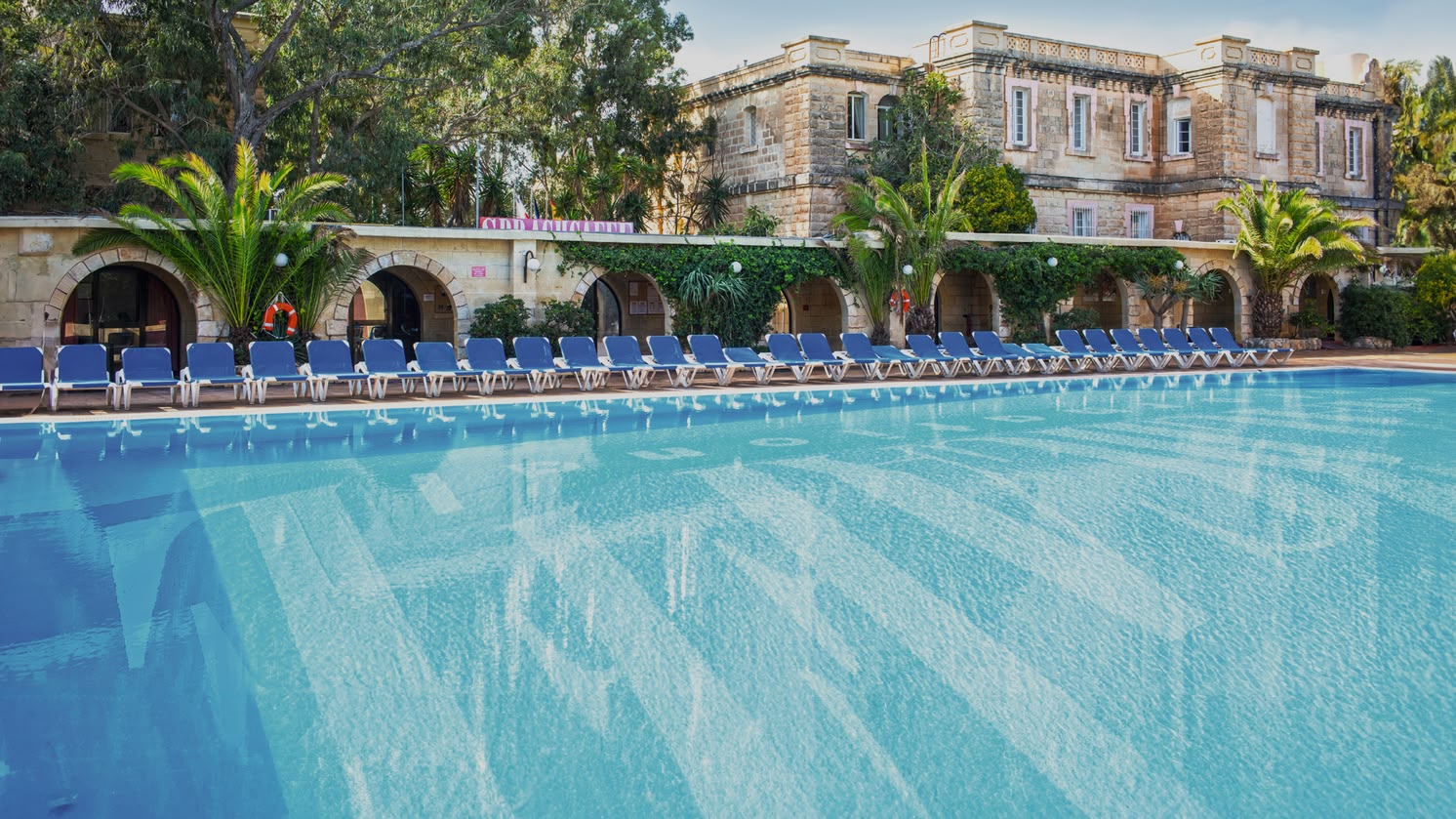 Large outdoor swimming pool surrounded by sun loungers and Mediterranean-style buildings at Sprachcaffe Malta.