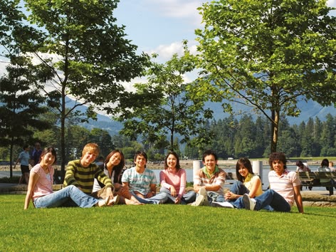 Groep studenten zittend op het gras in een park in Vancouver met bomen en bergen erachter.