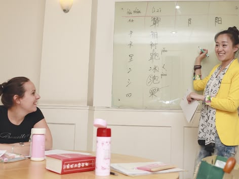 Leraar legt Chinese karakters uit op een whiteboard terwijl een leerling luistert en glimlacht.