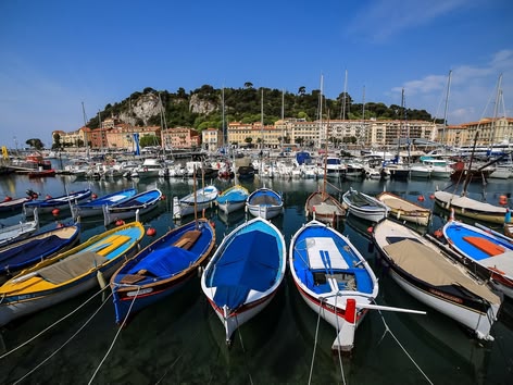 Kleurrijke boten aangemeerd in de haven van Nice, Frankrijk, met gebouwen op een heuvel en een jachthaven op de achtergrond.