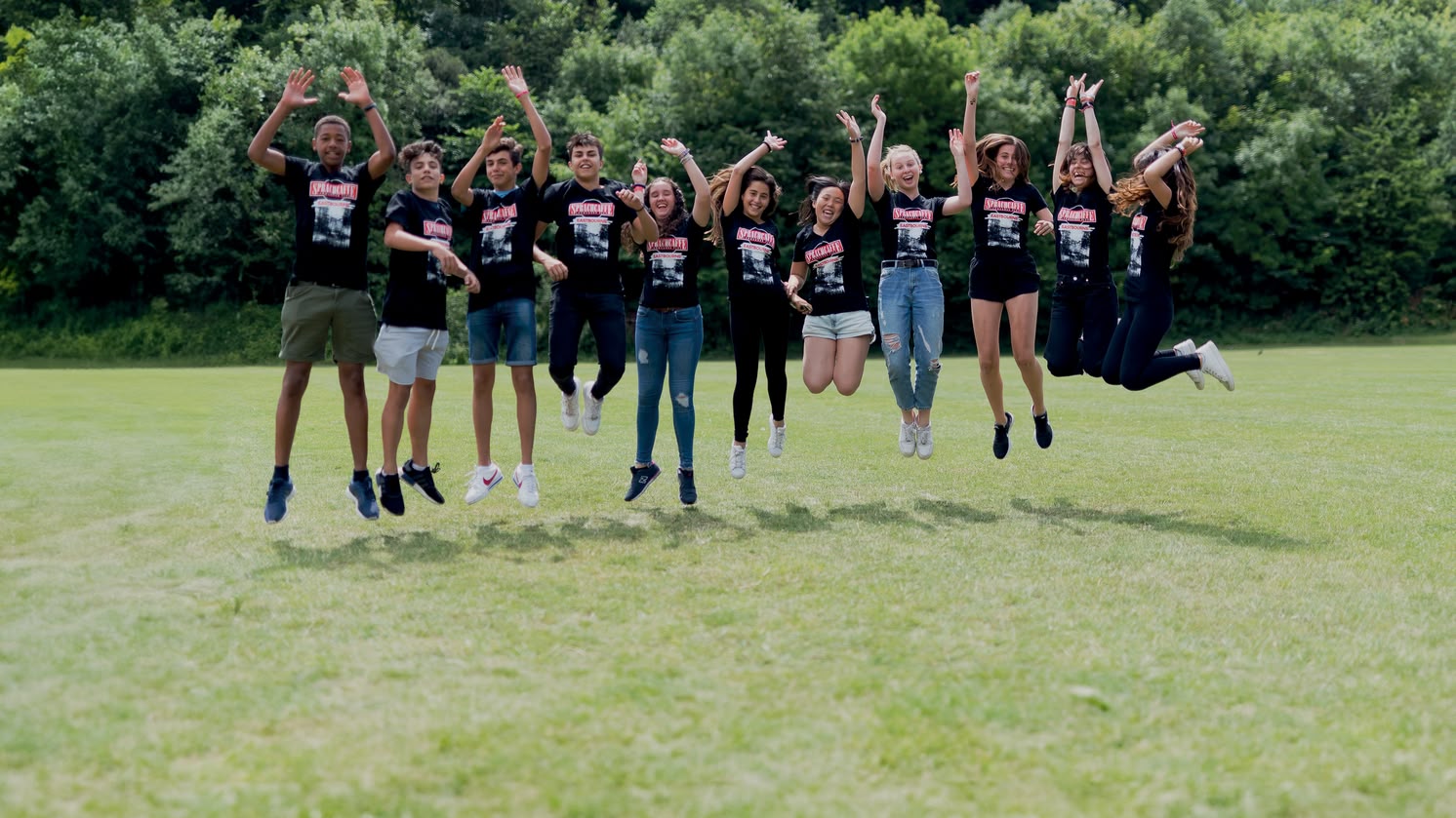 Sprachcaffe studenten in Engeland springen in de lucht op een groen veld, dragen bijpassende shirts en genieten van de buitenlucht.