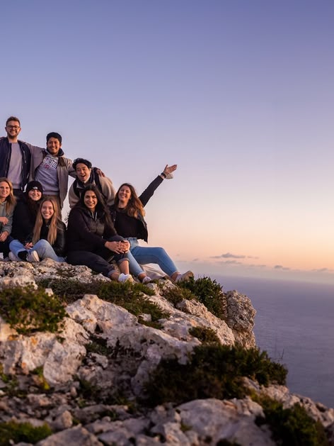 Groep jongvolwassenen op een rotsachtige klif in Malta bij zonsondergang, uitkijkend over zee en genietend van het uitzicht.