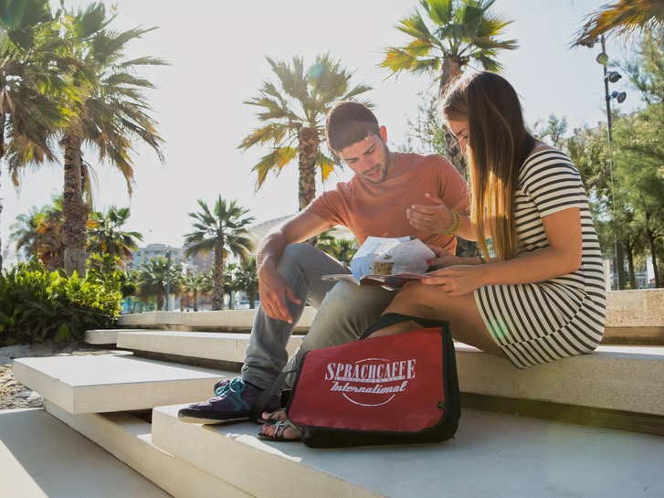 Twee studenten zitten aan zee in Spanje met een tas Sprachcaffe, omringd door palmbomen en moderne trappen.