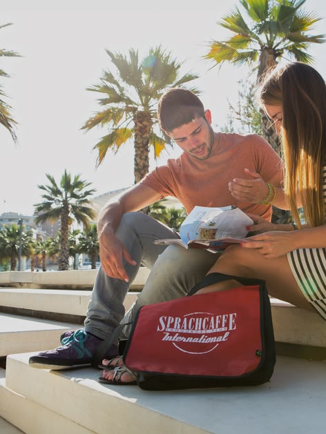 Twee Sprachcaffe studenten zitten aan het strand in Spanje een kaart te lezen met palmbomen op de achtergrond.